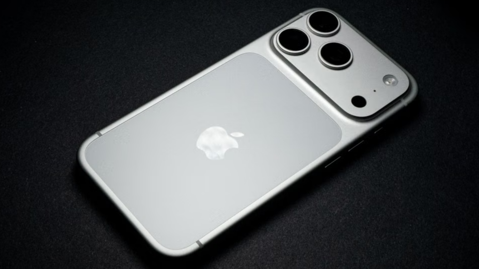 Apple May Raise Prices as Memory Chip Costs Climb. Will Consumers Keep Buying? The matte-finished back panel of a silver iPhone Pro model resting on a black surface, highlighting its triple-lens camera array and central Apple logo.