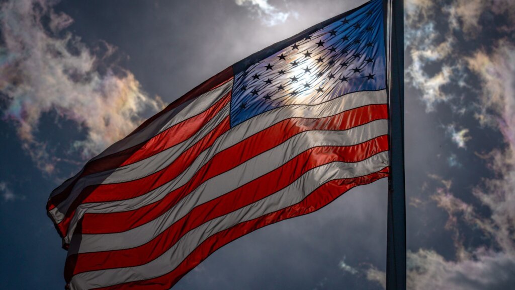 American flag on a pole waving against a dramatic cloudy sky with sunlight.