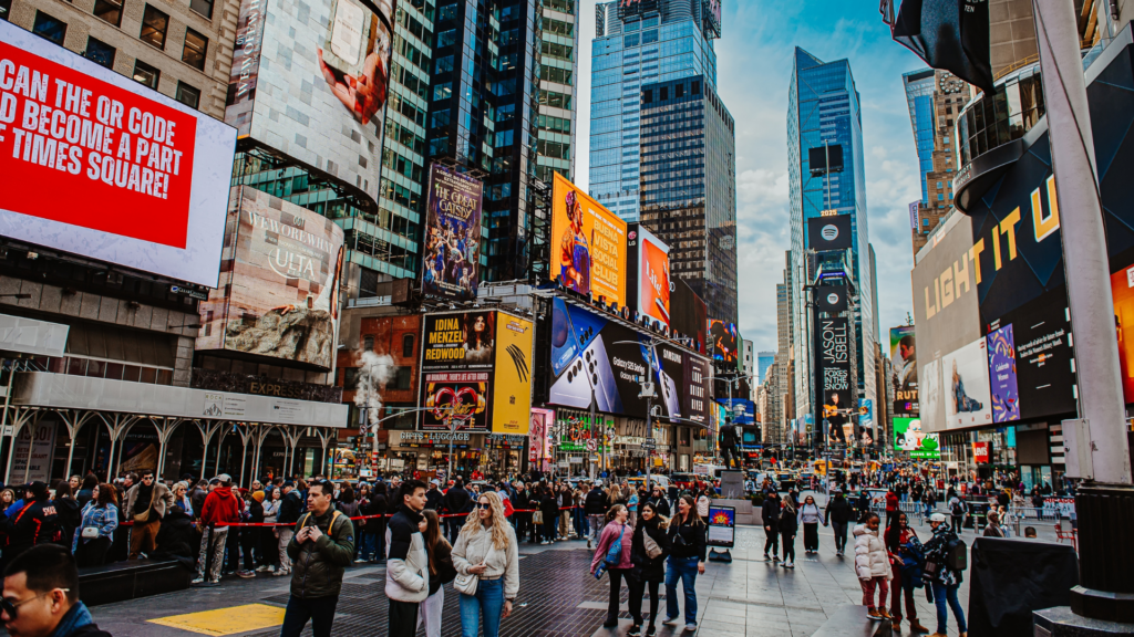A bustling, low-angle view of Times Square filled with pedestrians walking past towering skyscrapers covered in massive, glowing digital billboards and advertisements.