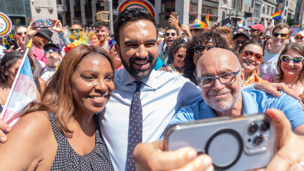 A close-up selfie taken on a smartphone showing Zohran Mamdani smiling broadly in a light blue shirt and tie, surrounded by enthusiastic supporters at a vibrant Pride event.