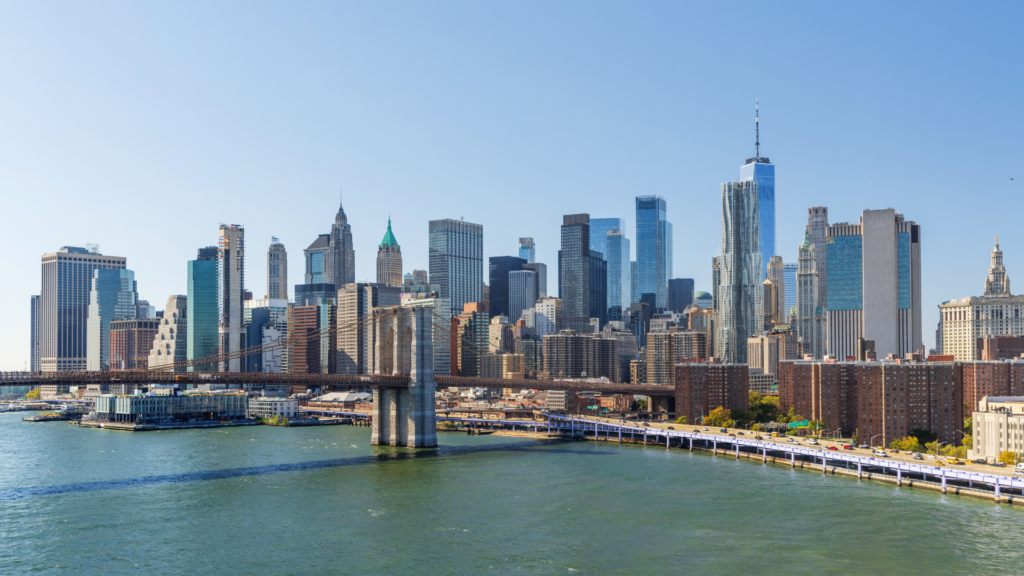 A wide, clear daytime shot of the Lower Manhattan skyline across the East River, with the iconic stone towers of the Brooklyn Bridge stretching across the foreground.