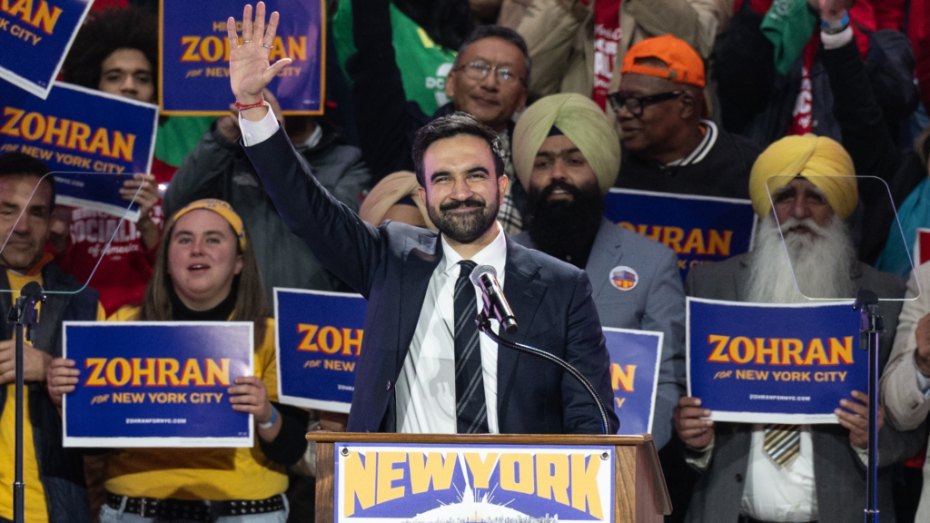 Mayor Zohran Mamdani stands at a wooden podium labeled New York Is Not For Sale, smiling and waving to a diverse crowd holding blue campaign signs that read Zohran for New York.