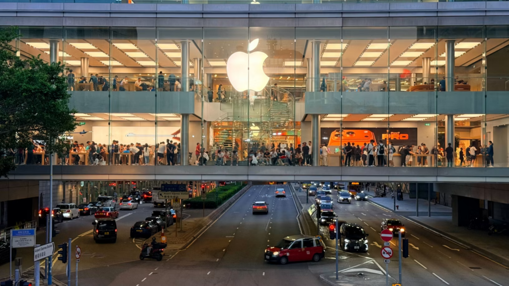 A wide, multi-level view of the illuminated glass facade of the Apple Store, spanning across an active city roadway.