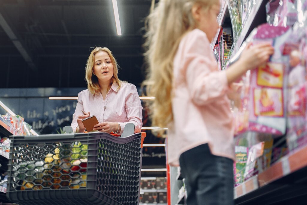 Woman reviews a shopping list as a child selects items from a supermarket shelf.