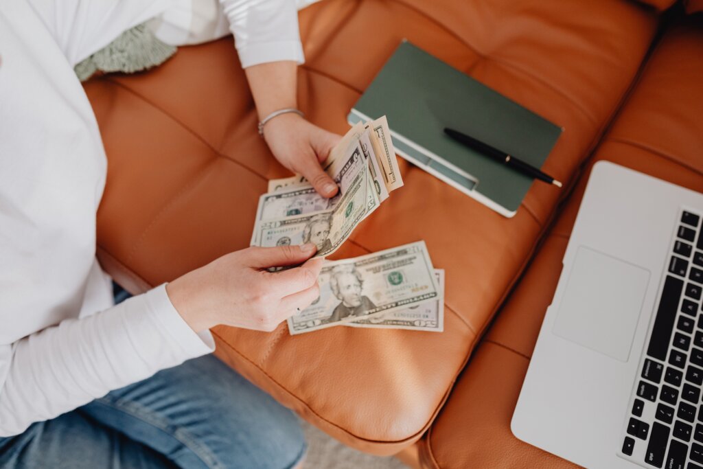 Person counting U.S. dollar bills beside a laptop and notebook.
