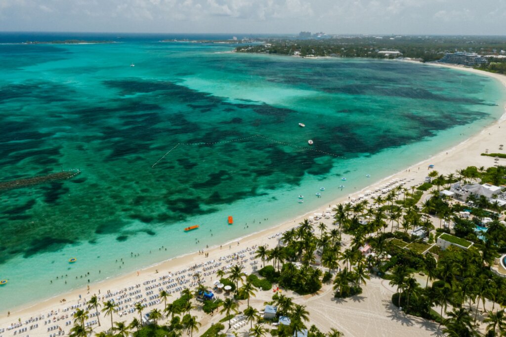 Aerial view of a white-sand beach and turquoise waters in the Bahamas, lined with palm trees and beachfront resorts.