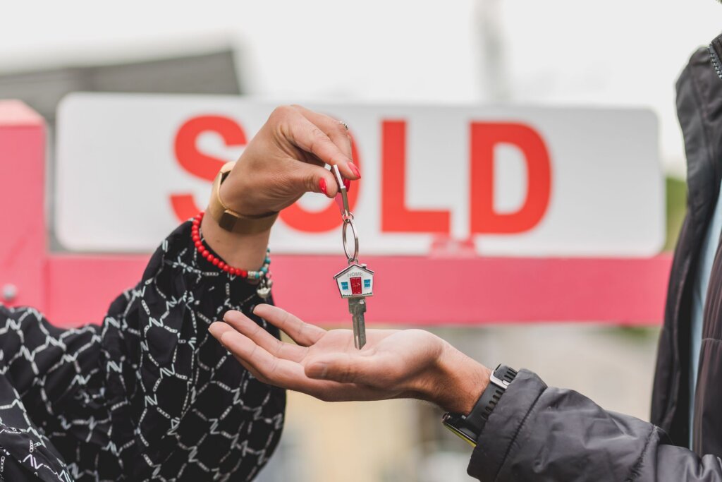 Person handing over a house key with a home-shaped keychain in front of a sold real estate sign.