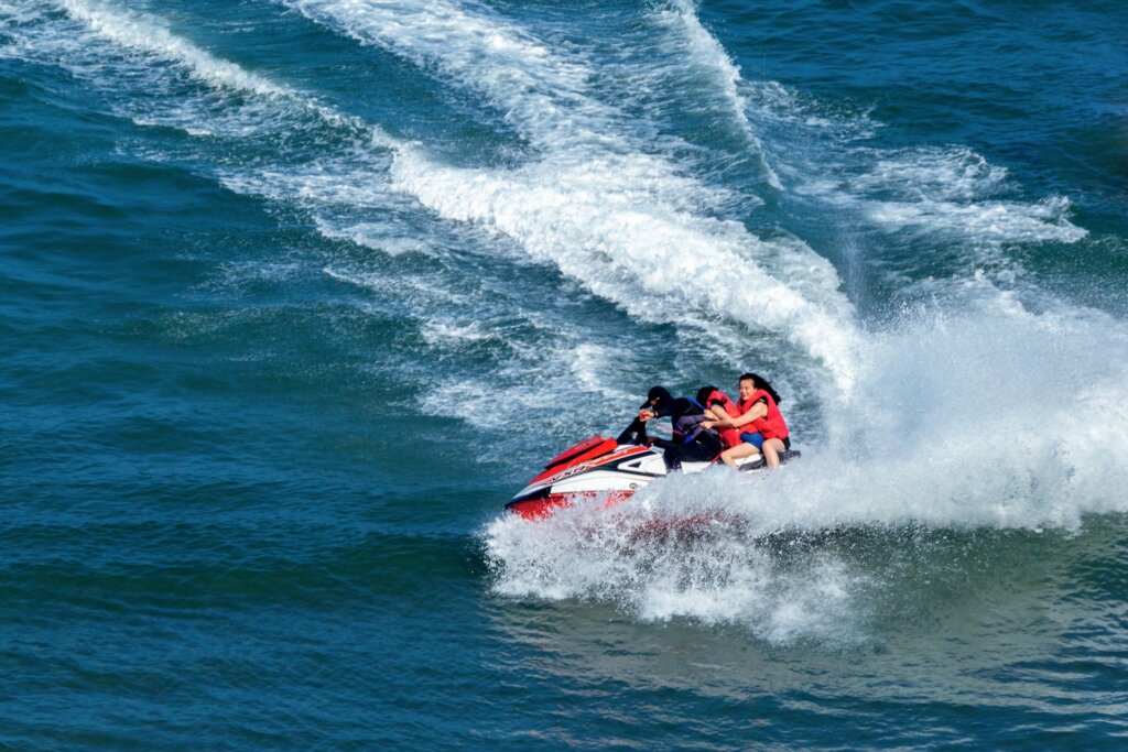 Two guests wearing life jackets ride with a jet ski operator across open water, leaving a foamy wake behind them.