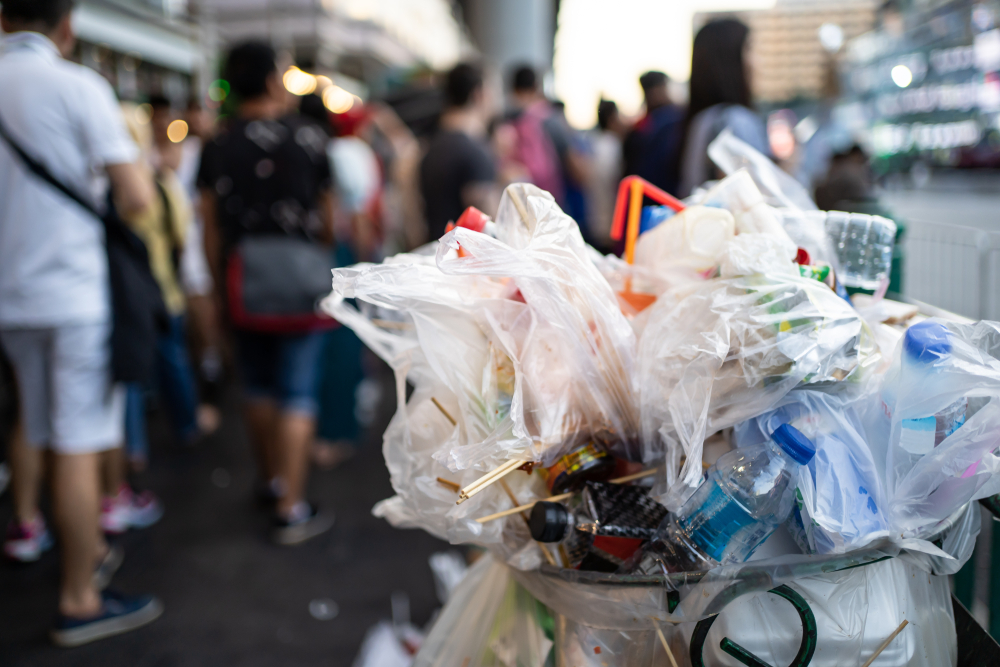 Overflowing trash bin filled with plastic waste on a busy city street crowded with pedestrians.