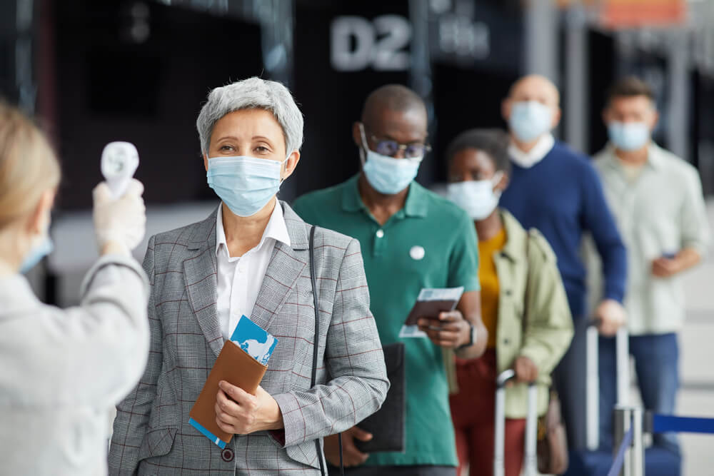Masked travelers wait in line at an airport as a staff member conducts temperature screening.