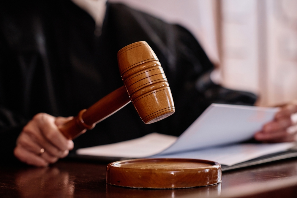 A judge holds a gavel above the sounding block while reviewing documents at a courtroom bench.