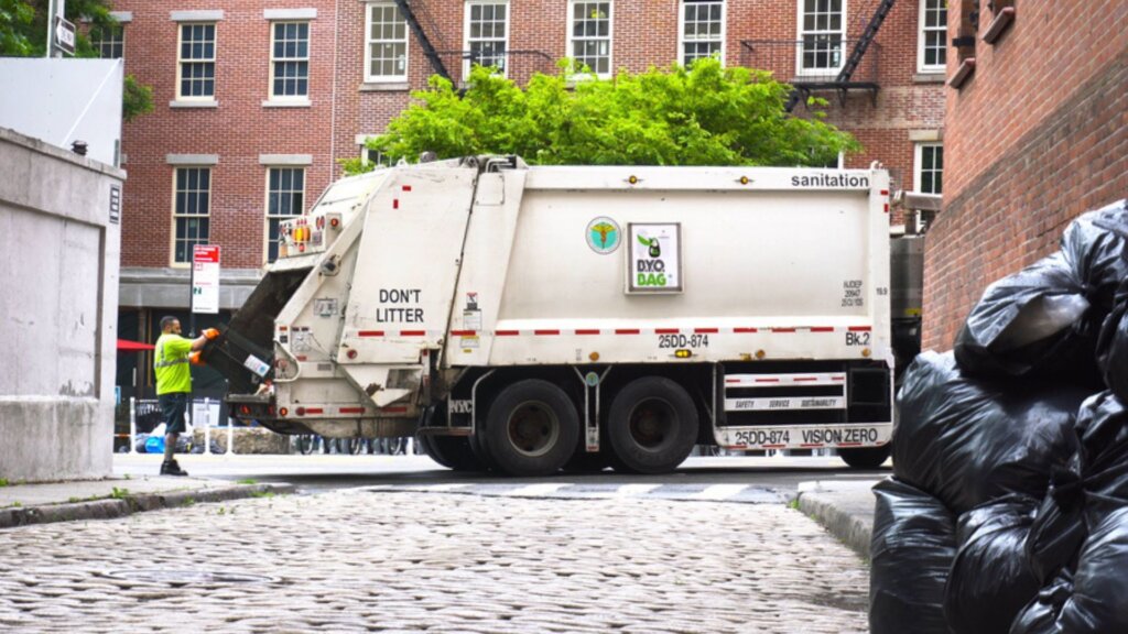 New York City Department of Sanitation truck collecting trash on a cobblestone street lined with garbage bags.