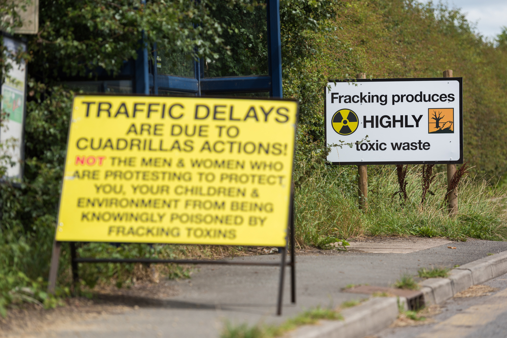Roadside protest signs criticizing fracking, including warnings about toxic waste and traffic delays near a drilling site.