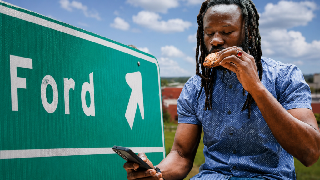 Man using a phone beside a Ford road sign while eating a cookie.