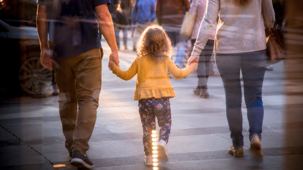 Parents walk hand in hand with their young child along a busy city sidewalk at sunset.