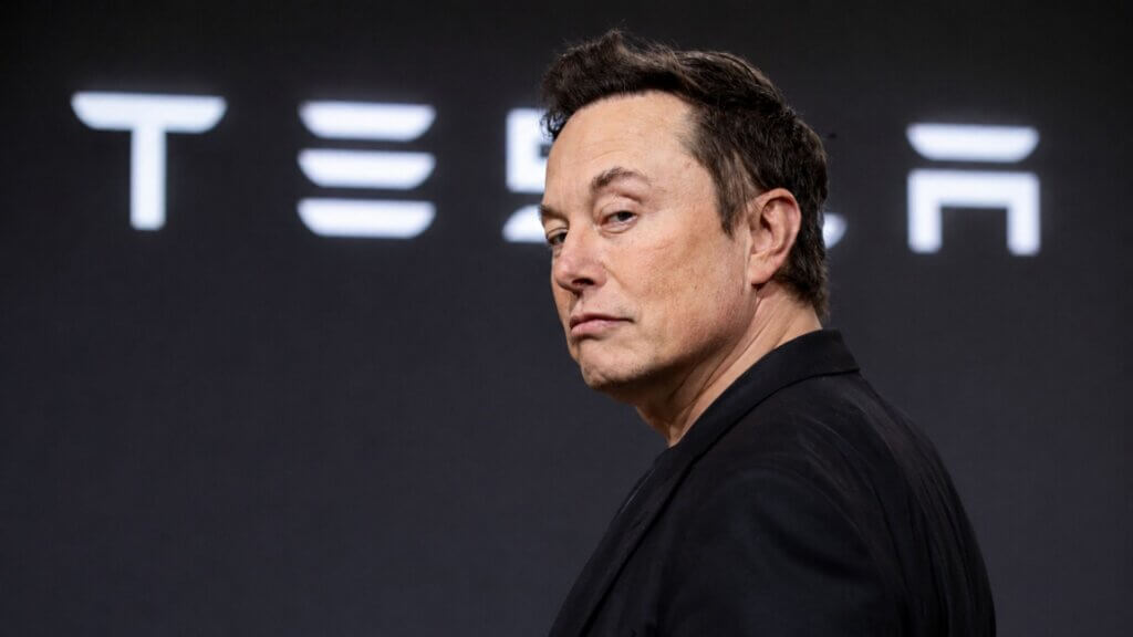 Elon Musk stands in front of a blurred Tesla logo backdrop while looking over his shoulder at the camera during a Tesla event.