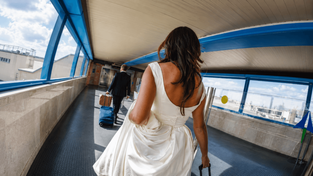 An over-the-shoulder shot of a bride in her wedding dress pulling a suitcase through an airport terminal walkway following the groom.