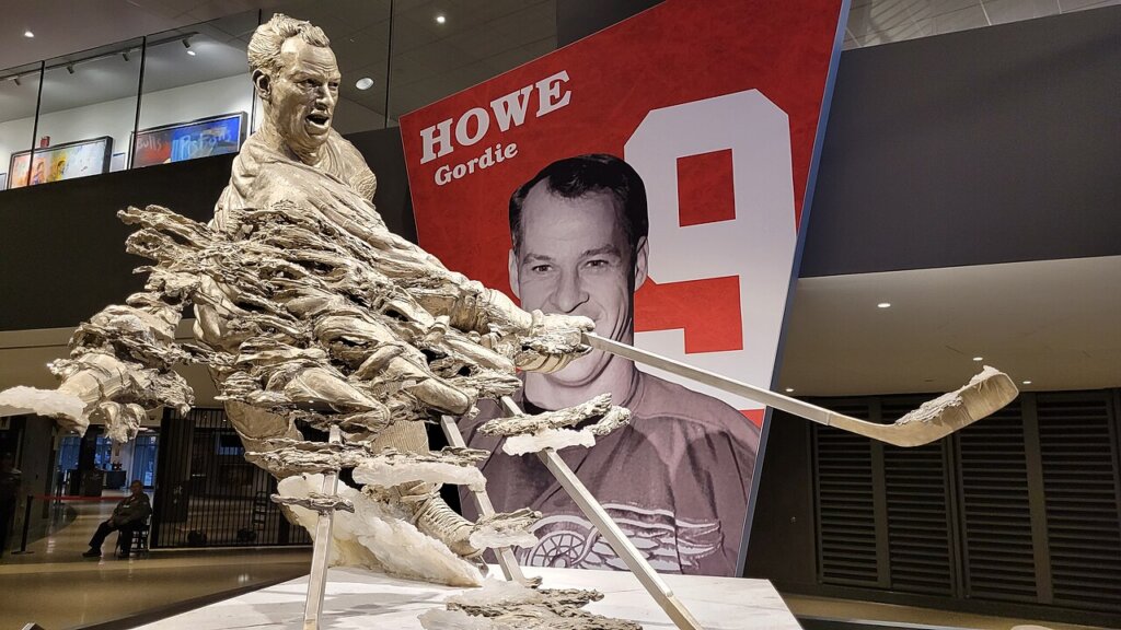 Statue of Gordie Howe displayed inside Little Caesars Arena with a large portrait of the hockey legend in the background.