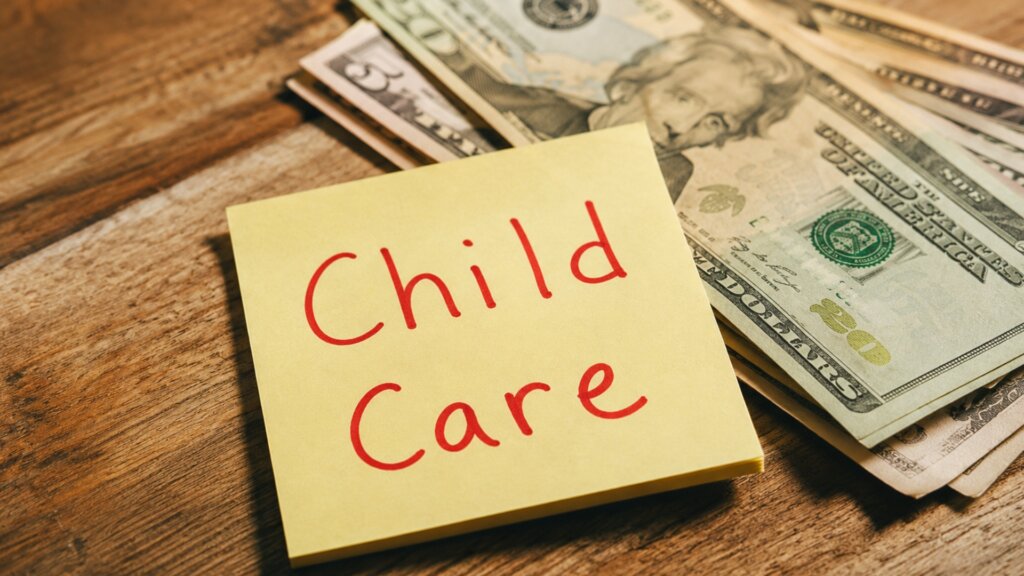 A stack of U.S. dollar bills sits beside a yellow sticky note that reads "Child Care" in red handwriting.