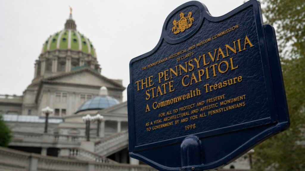 Historic marker for the Pennsylvania State Capitol stands in the foreground with the capitol building and its green dome blurred in the background.