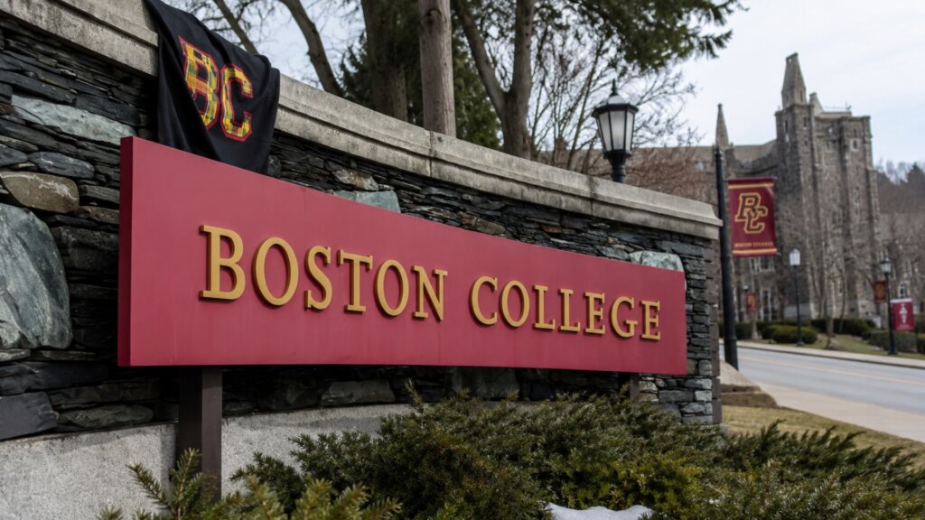 Stone entrance sign reading "BOSTON COLLEGE" with the campus and BC banners visible in the background.