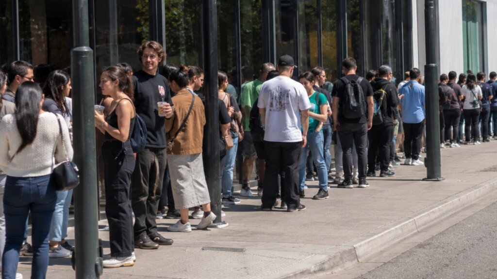 A long line of people waits outside a modern building on a sunny day, with the queue stretching along the sidewalk.