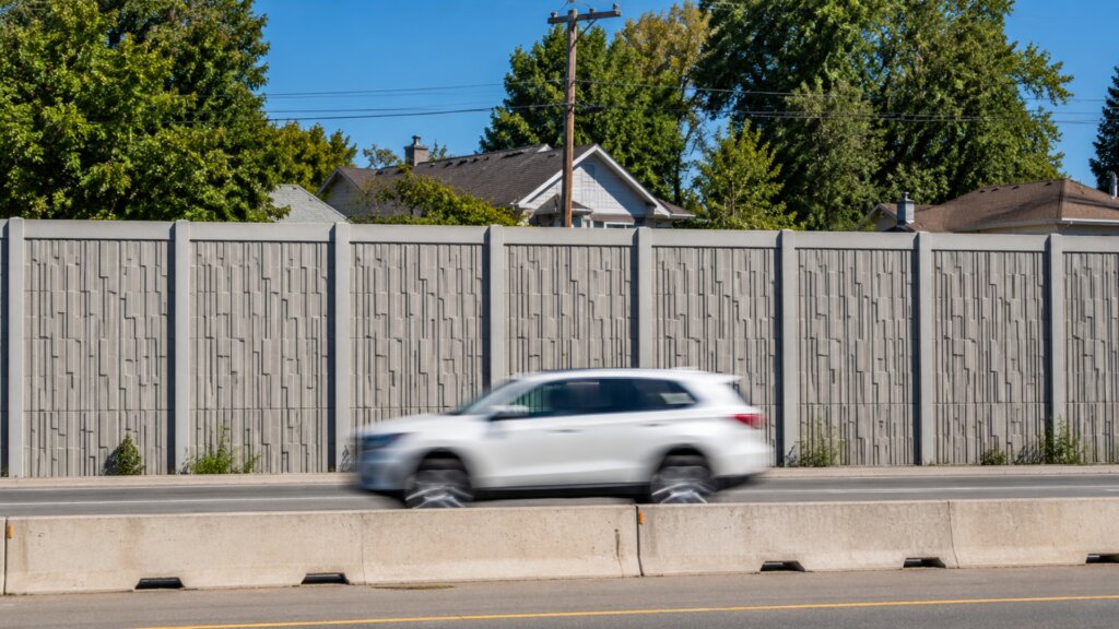 White SUV driving past a tall concrete sound barrier with residential homes and trees visible behind it.
