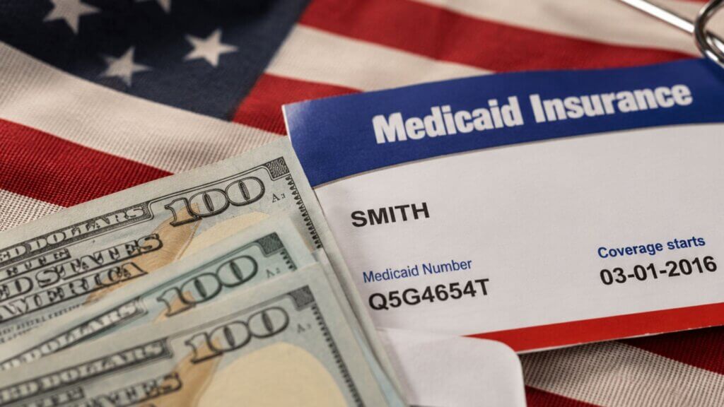 A Medicaid insurance card labeled "SMITH" rests beside U.S. one hundred dollar bills on an American flag.