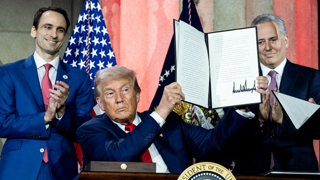 Donald Trump sitting at a desk holding up a signed official document while two men in suits applaud behind him.