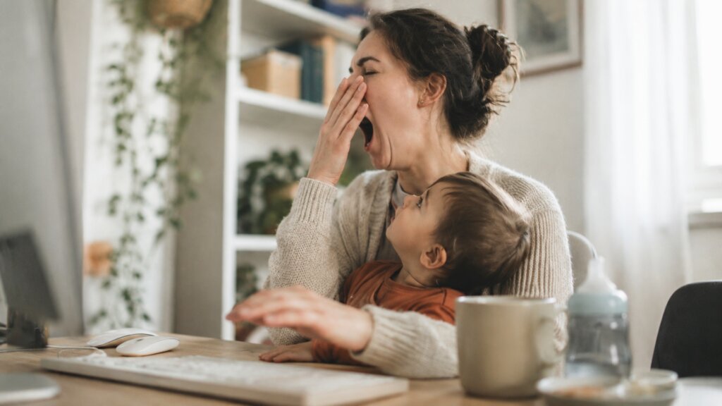 A tired parent yawns while sitting at a computer with a young child on their lap in a home office.