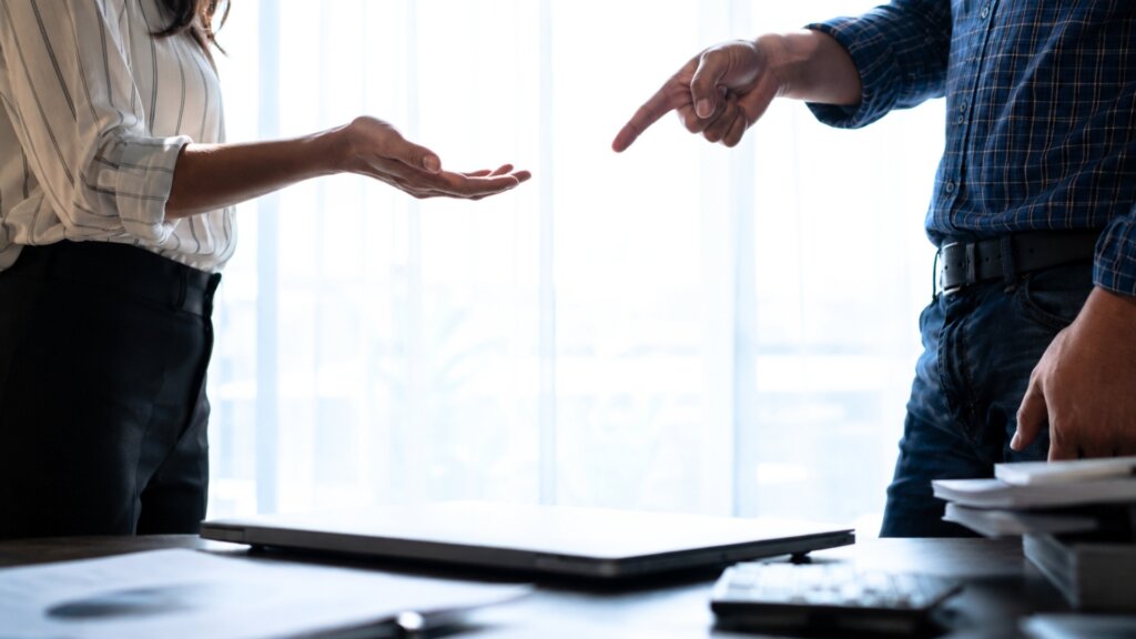 Two people stand across from each other in an office as one points a finger while the other holds out an open hand during a tense discussion.