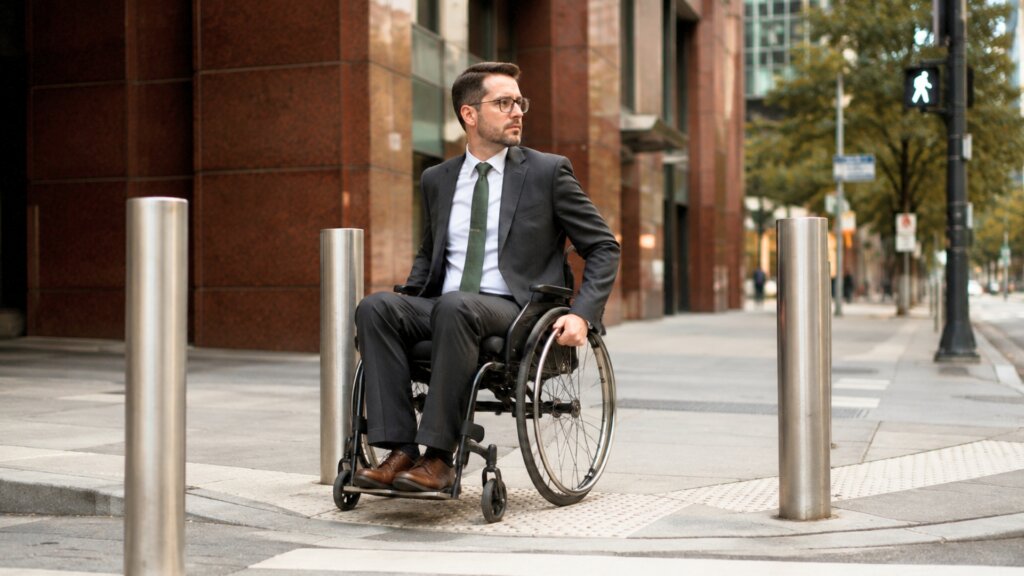 A man using a wheelchair waits at an accessible street crossing between metal bollards outside office buildings in an urban setting. The image highlights accessible infrastructure and mobility in a public space.