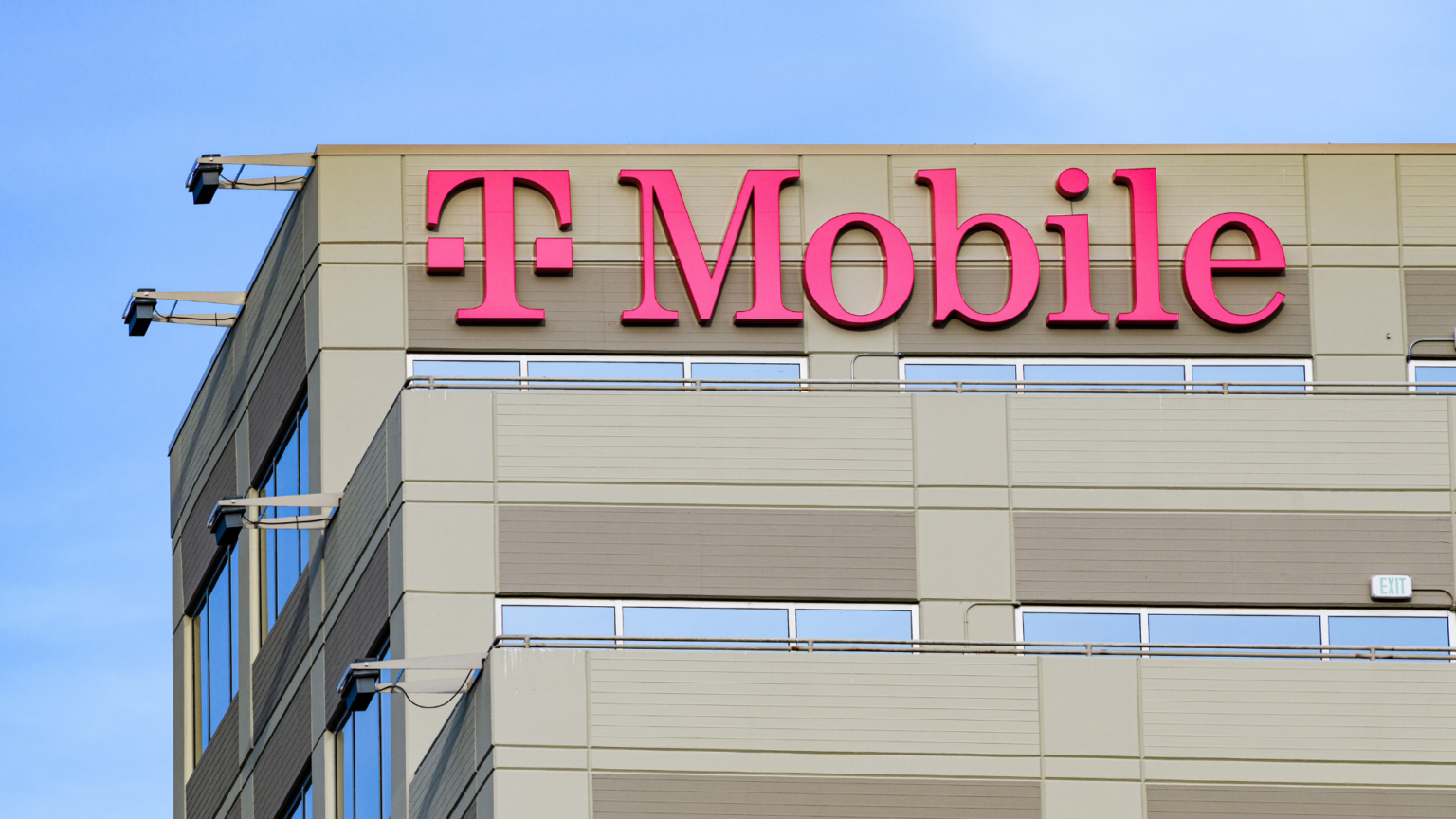 A bright pink T-Mobile sign on the exterior of a grey commercial building against a clear blue sky.