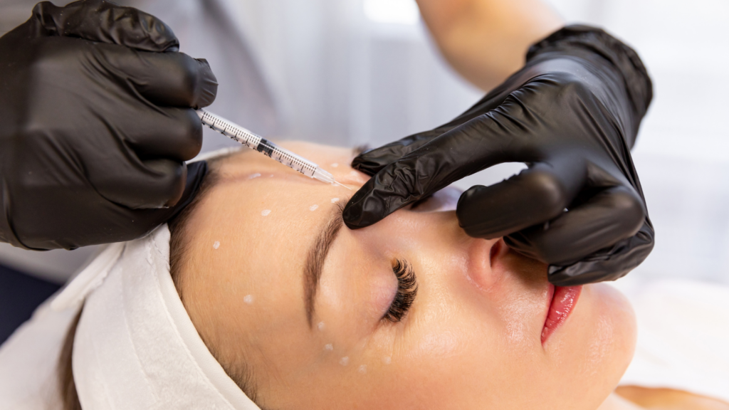A close-up of a gloved hand administering an injection into a woman's forehead, which is marked with white dots.