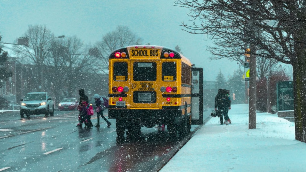 A yellow school bus stopped on a road during a heavy snowstorm with people walking nearby.