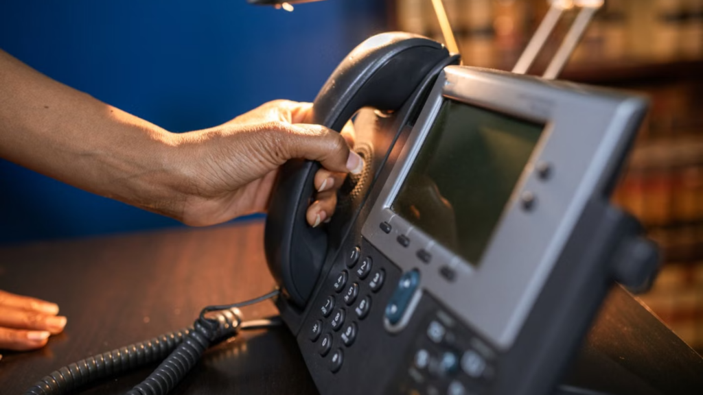 A close-up of a person's hand picking up the receiver of a modern grey multi-line office telephone.