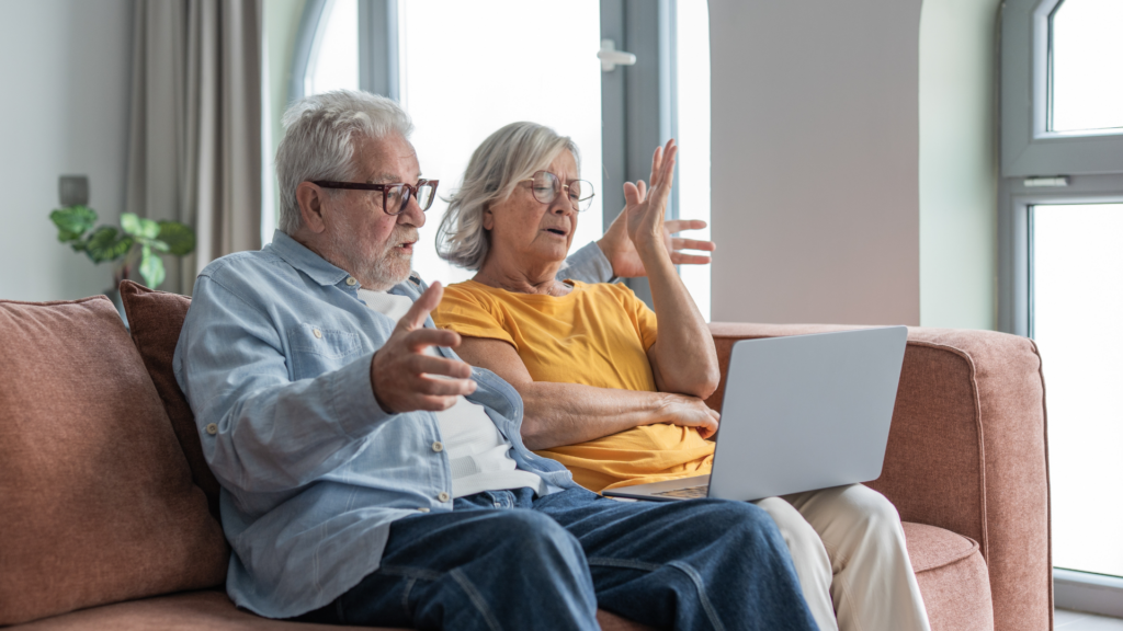 An older man and woman sitting on a couch, looking confused and frustrated at a laptop computer.