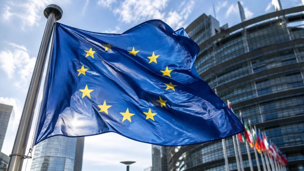 A European Union flag waves in front of a modern government building under a blue sky.