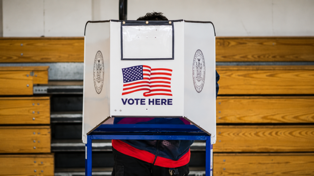 A voter cast their ballot inside a white voting booth featuring an American flag graphic and "Vote Here" text.