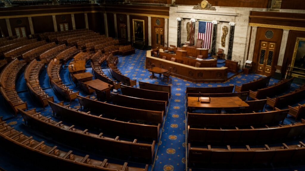 Wide view of the mostly empty U.S. House chamber with rows of wooden seats facing the speaker's podium beneath a large American flag.