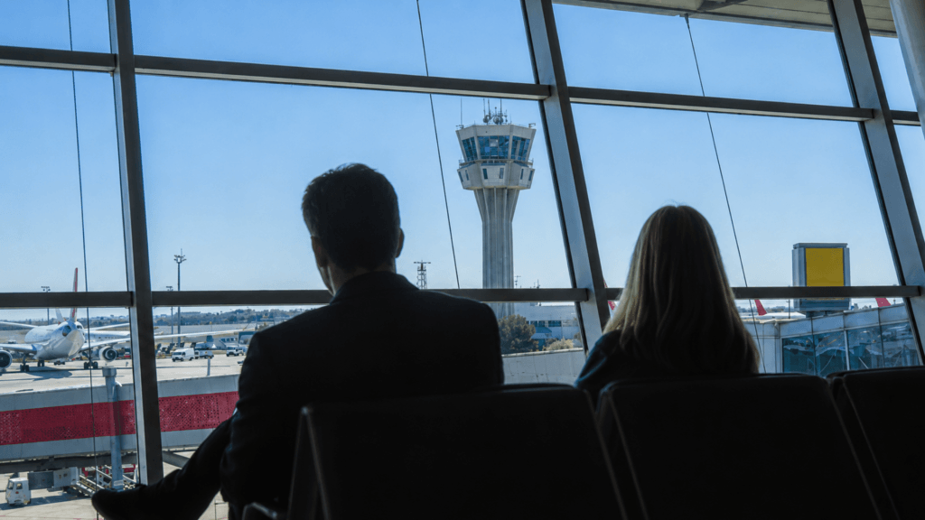 The silhouette of a couple waiting inside an airport terminal while looking through large windows at the control tower and airfield.