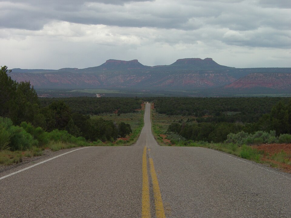 A two-lane road stretches toward the mesas of Bears Ears National Monument in southern Utah beneath a cloudy sky.