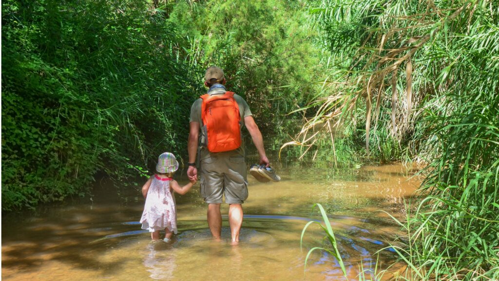 A father and his young daughter walking through a river surrounded by North Dakota’s scenic landscape.