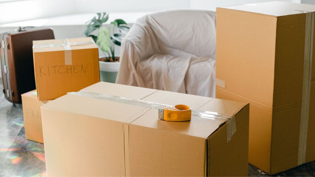 Cardboard moving boxes, one labeled "KITCHEN" in marker, stacked in a living room with a sheet-covered armchair, a potted plant, a suitcase, and a tape gun sitting on top of a taped box.