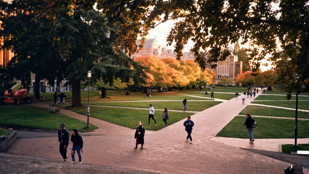 Students walking on paved paths across a college quad lined with autumn trees, with Gothic-style campus buildings in the background.