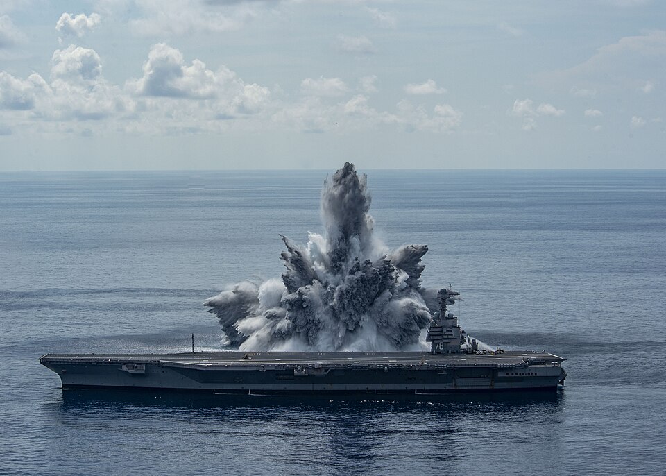 Controlled explosion erupts beside a U.S. Navy aircraft carrier during a Full Ship Shock Trial.