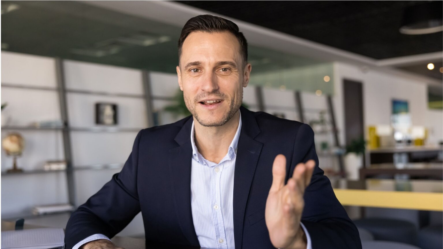 Businessman in a navy suit sitting at a desk, speaking and gesturing with one hand as if talking to someone.