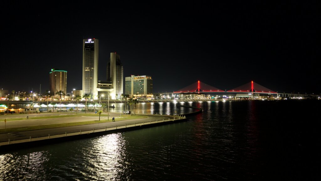 Nighttime view of the Corpus Christi, Texas, waterfront skyline, with illuminated downtown buildings and the Harbor Bridge reflected in Corpus Christi Bay.