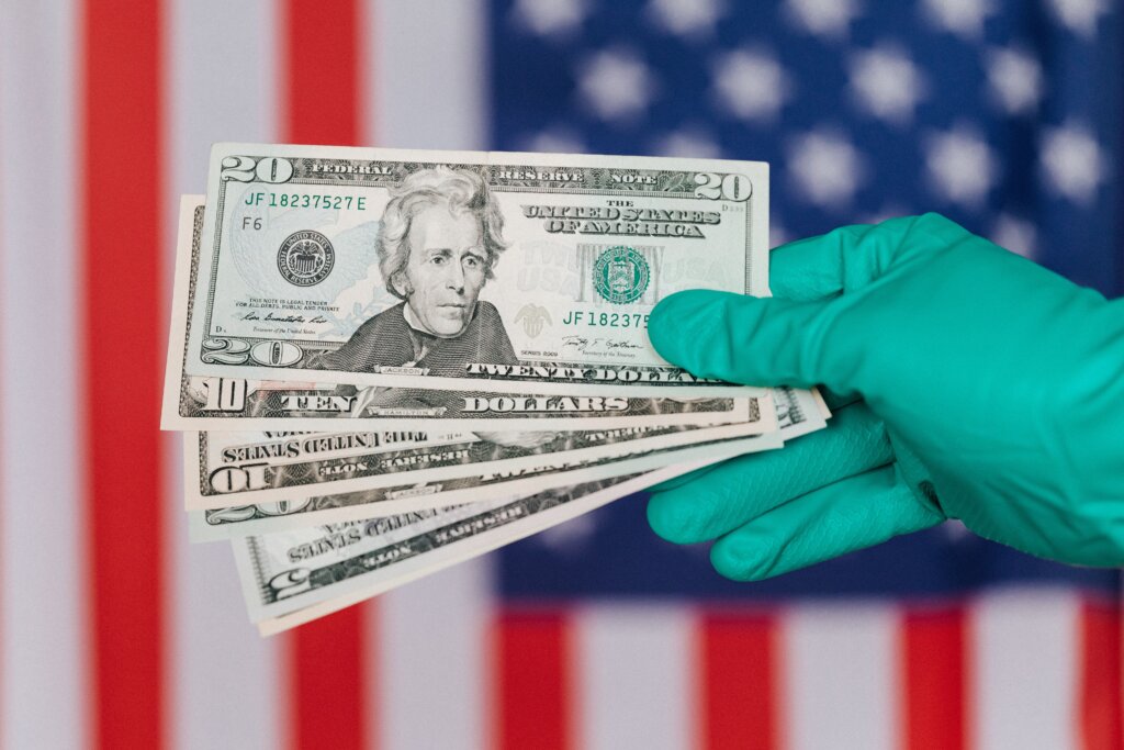 A gloved hand holds U.S. dollar bills against an American flag background, illustrating the financial costs associated with healthcare and medical expenses.