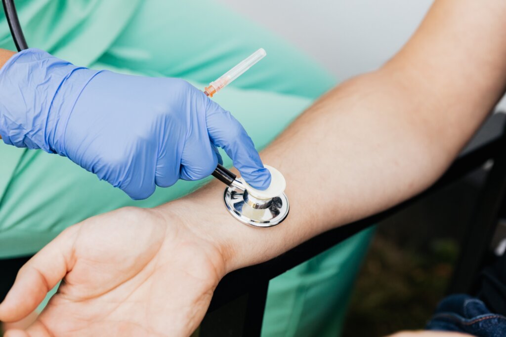 A healthcare worker wearing blue gloves uses a stethoscope to examine a patient's arm during a routine medical checkup.